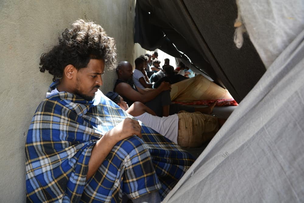 Refugees outside try to protect themselves from the sun by leaning back on the walls of the buildings in Zintan detention centre. Libya, June 2019. ©JÉRÔME TUBIANA/MSF