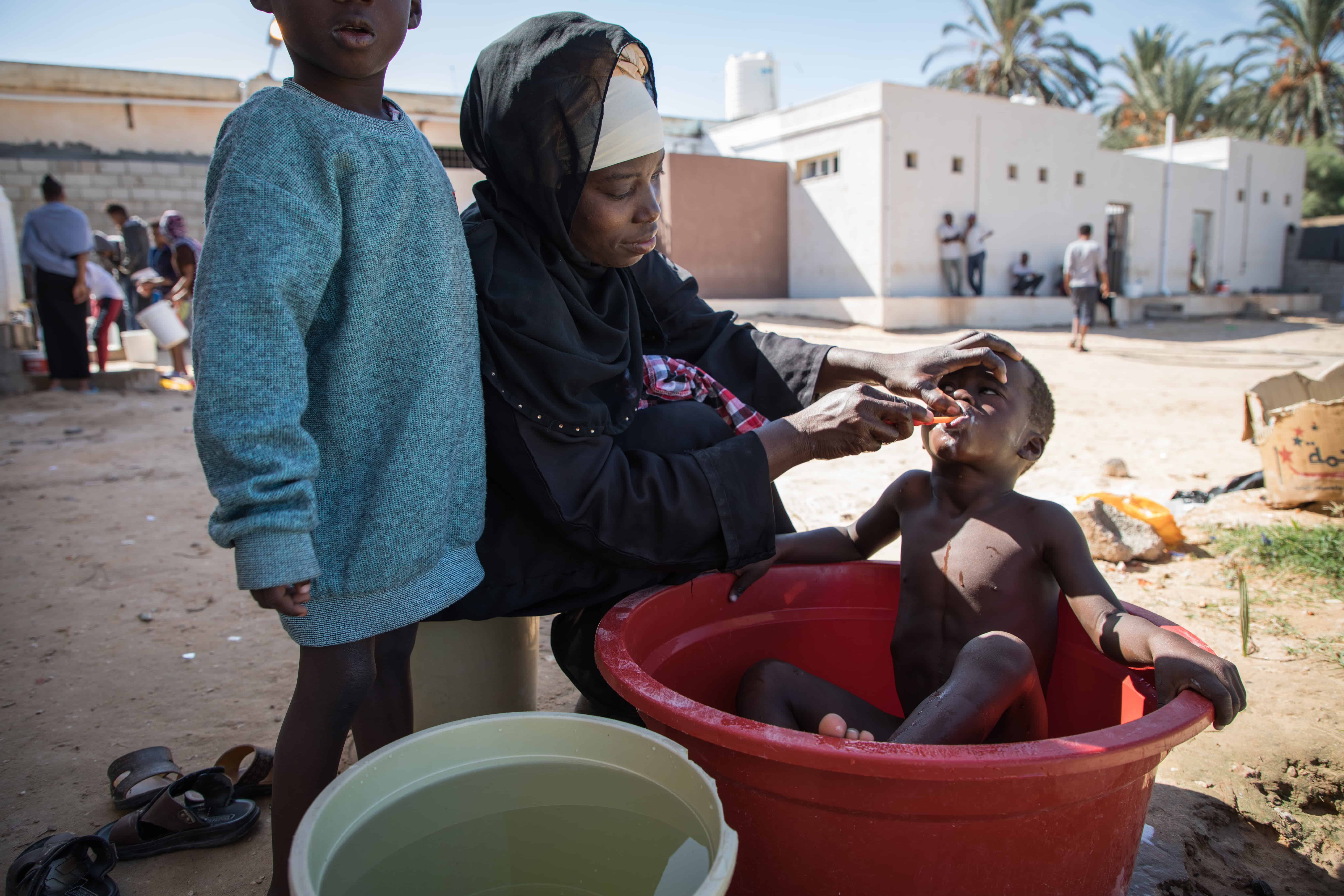 A mother brushes her child's teeth while giving him a bath within the confines of the Souq Al-Khamis detention centre. Khoms, Libya, October 2019. © AURELIE BAUMEL/MSF